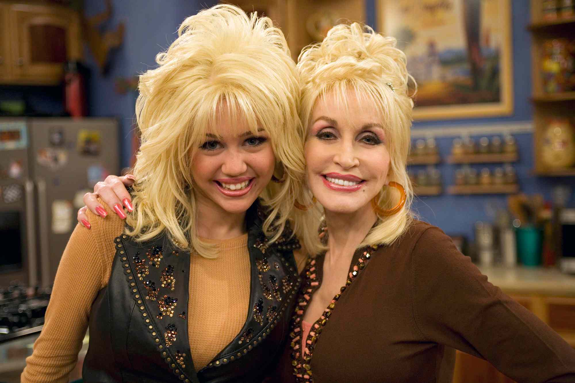 Miley Cyrus e Dolly Parton sorrindo, com cabelos loiros volumosos e maquiagem marcante, em um cenário de cozinha com armários e prateleiras