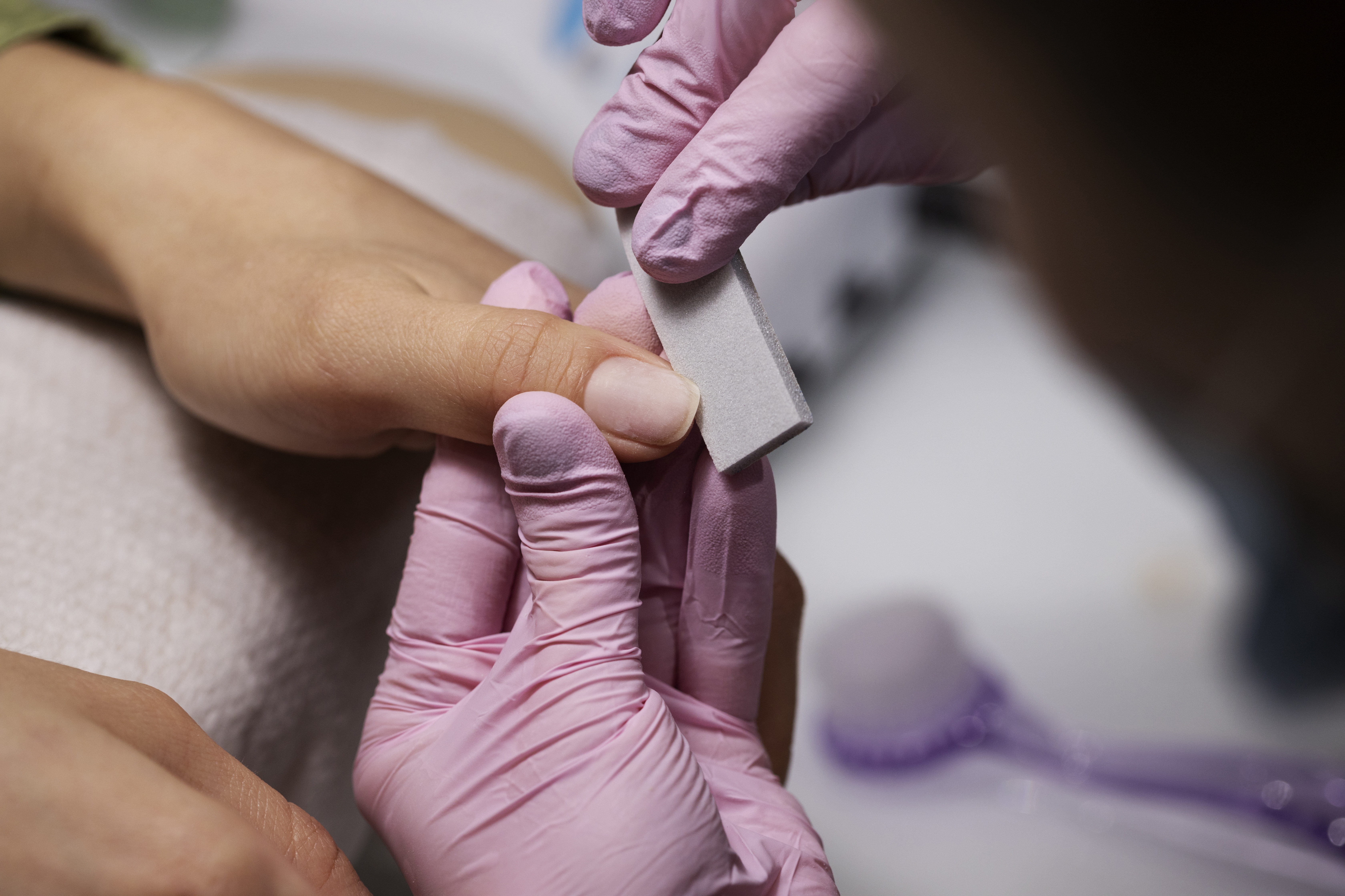 M&atilde;os de uma manicure com luvas rosa lixando a unha de uma cliente, que tem a m&atilde;o apoiada em uma toalha clara