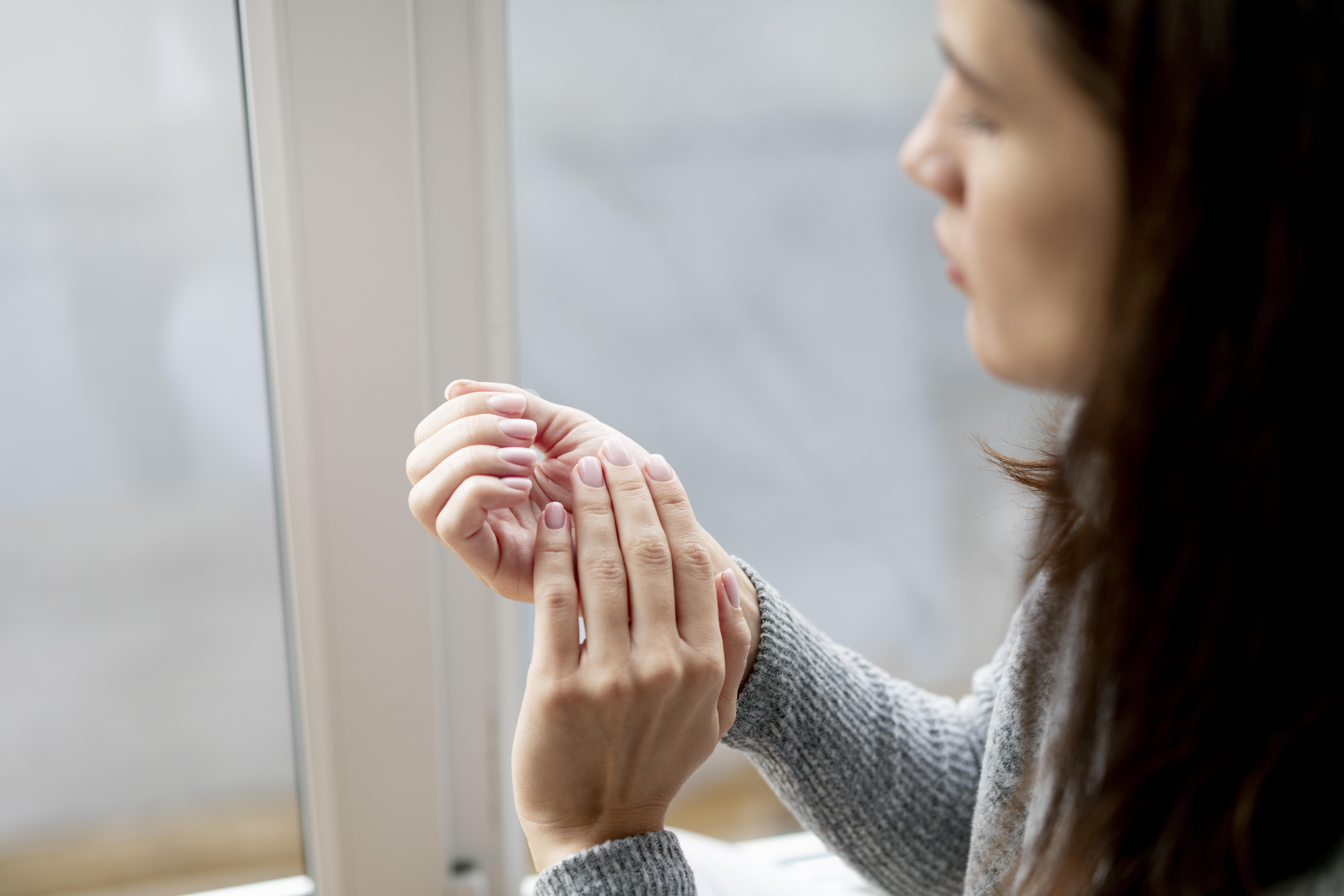 Mulher de cabelos escuros e su&eacute;ter cinza massageia o pulso esquerdo com a m&atilde;o direita, com express&atilde;o de dor ou desconforto, em ambiente interno com janela ao fundo