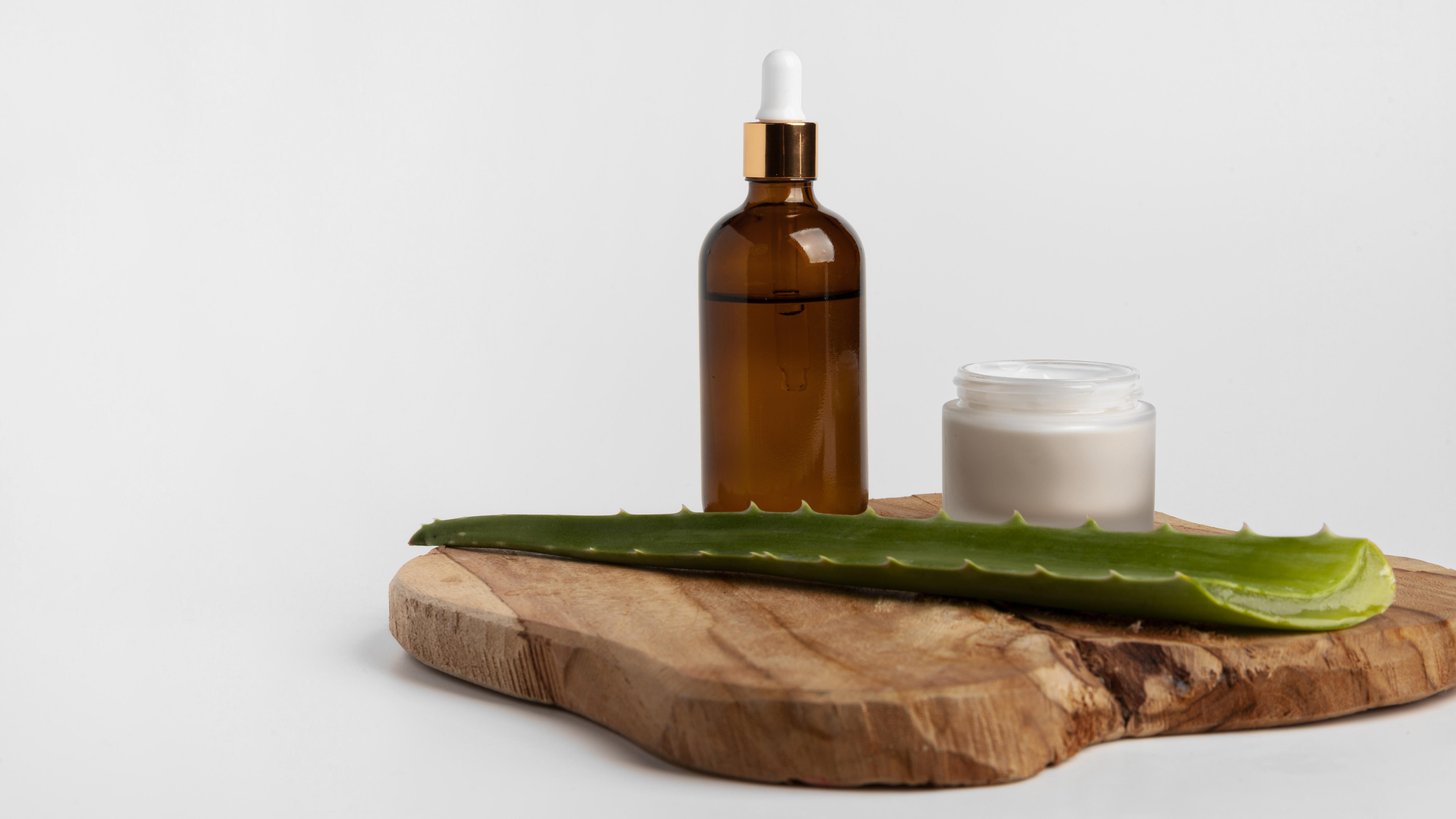 Uma folha de aloe vera verde com espinhos nas bordas, um frasco conta-gotas &acirc;mbar e um pote de creme branco sobre uma t&aacute;bua de madeira r&uacute;stica, em fundo branco