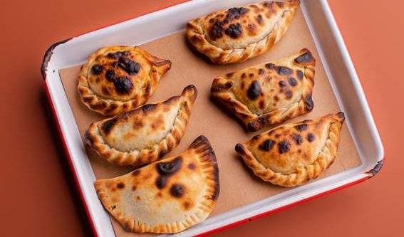 Seis empanadas assadas, com massa dourada e manchas escuras de cozimento, dispostas sobre papel manteiga em uma assadeira branca com borda vermelha, em fundo marrom-avermelhado