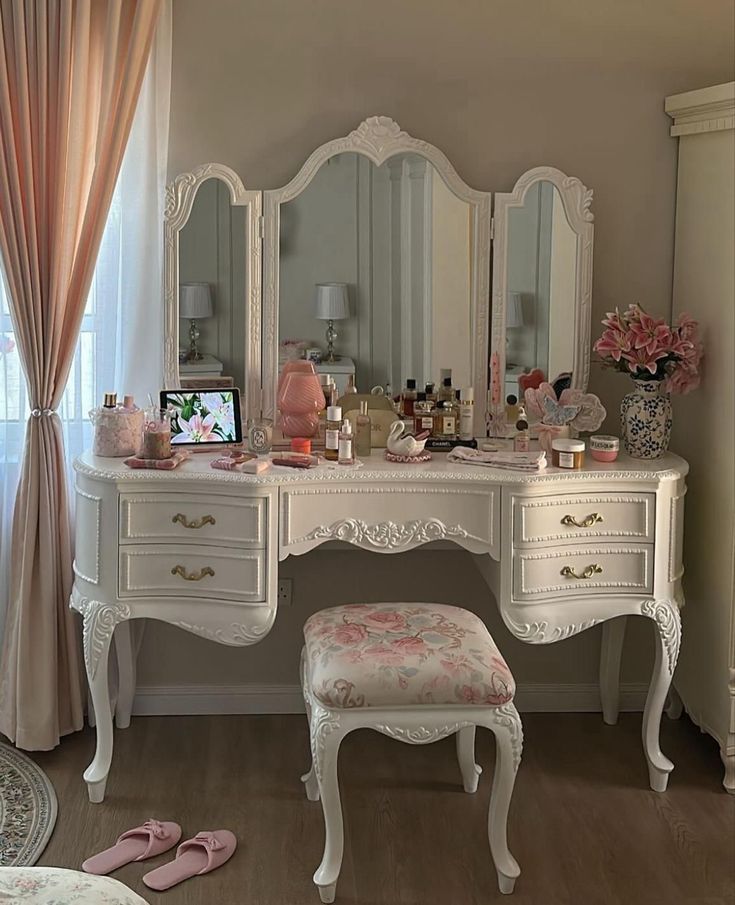 Penteadeira branca ornamentada com espelho triplo, repleta de maquiagens, perfumes e um tablet exibindo flores. Um banquinho estofado com estampa floral rosa e chinelos cor-de-rosa no ch&atilde;o completam o ambiente. Cortinas claras e rosadas ao lado.