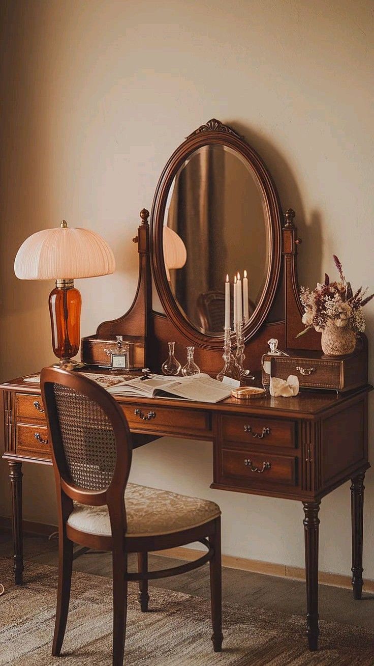 Penteadeira de madeira escura com espelho oval, gavetas e objetos de decora&ccedil;&atilde;o. H&aacute; um abajur com c&uacute;pula plissada, velas acesas em um casti&ccedil;al de vidro, frascos de perfume, um livro aberto e um vaso de flores secas. Uma cadeira de madeira com encosto de palha e assento estofado est&aacute; &agrave; frente da penteadeira