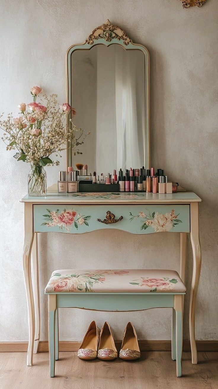 Penteadeira vintage azul-claro com detalhes florais e espelho dourado ornamentado. Sobre ela, um vaso com rosas cor-de-rosa e maquiagens diversas. Abaixo, um banco estofado com o mesmo padr&atilde;o floral e tr&ecirc;s pares de sapatilhas bege no ch&atilde;o de madeira
