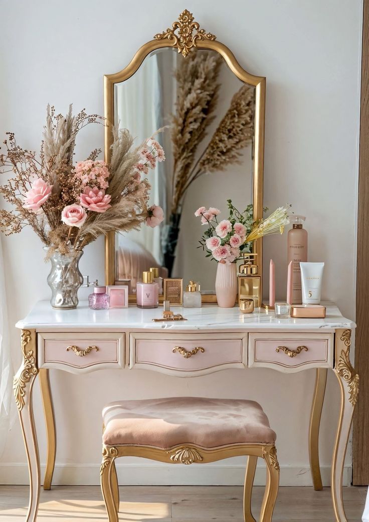 Penteadeira estilo proven&ccedil;al com espelho dourado ornamentado, tampo de m&aacute;rmore branco e gavetas rosa claro com puxadores dourados. Sobre o tampo, vasos com flores e folhagens secas em tons de rosa e bege, al&eacute;m de frascos de perfume e cosm&eacute;ticos. &Agrave; frente, um banco estofado em veludo rosa.
