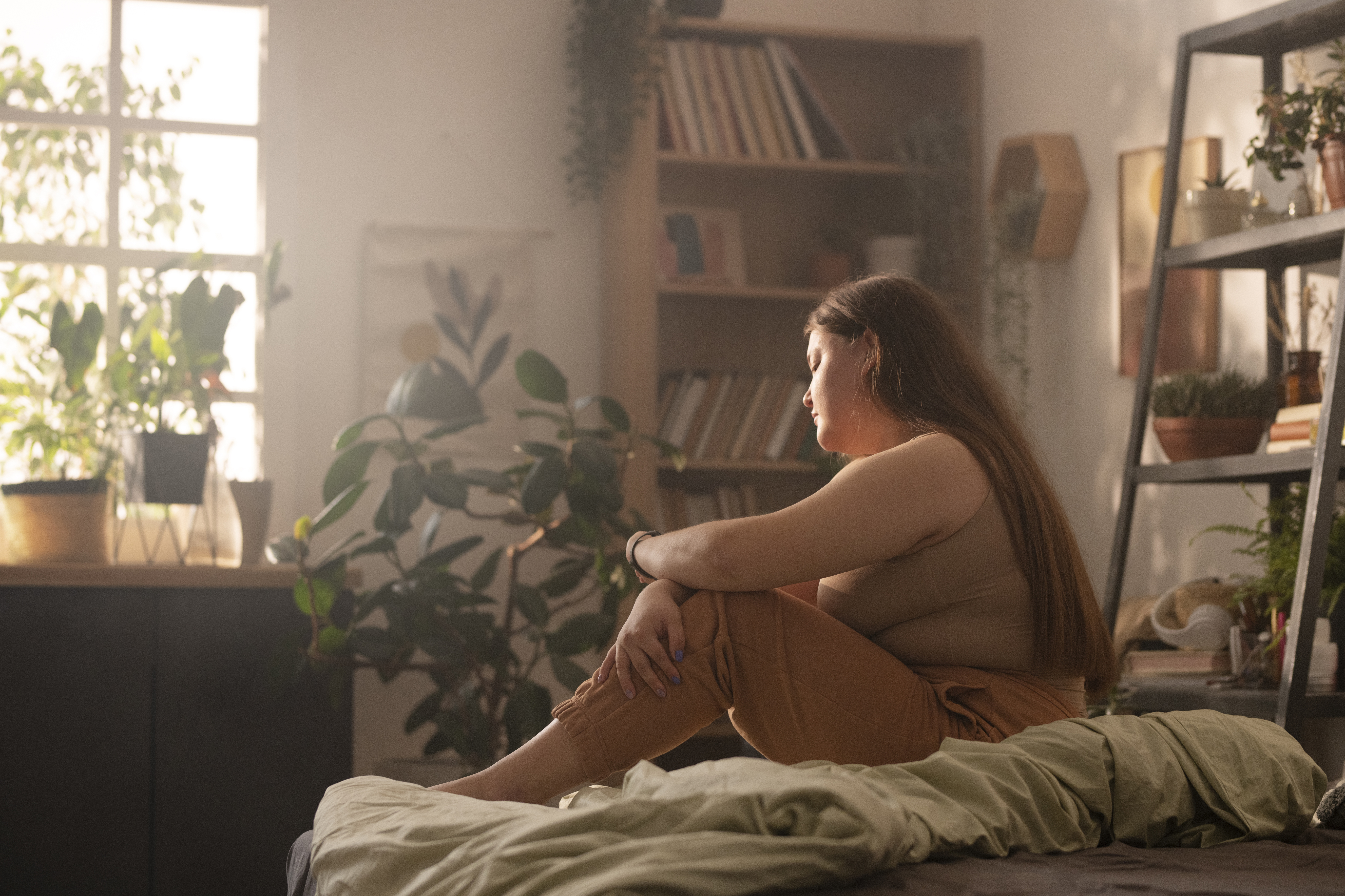 Mulher de cabelos castanhos e compridos, vestindo top e cal&ccedil;a marrons, sentada na cama com as pernas dobradas, olhando para a esquerda. O quarto tem luz suave, plantas e estantes com livros