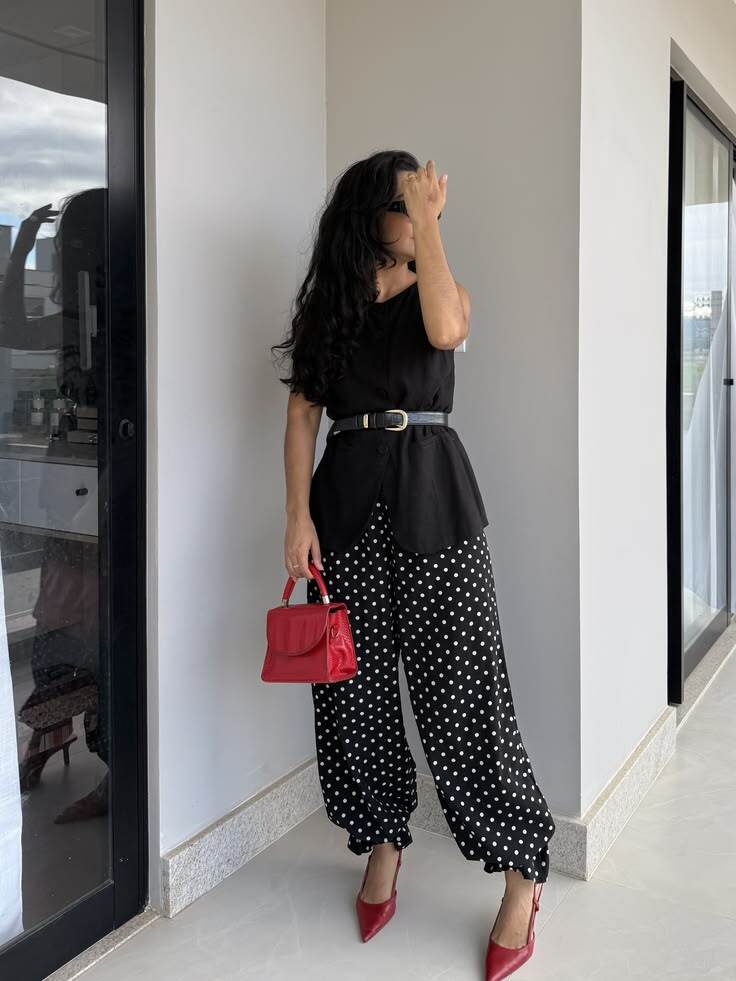 Mulher de cabelos escuros e &oacute;culos de sol, com blusa preta, cinto preto, cal&ccedil;a preta de bolinhas brancas, sapatos vermelhos de salto e bolsa vermelha, posando em um ambiente interno com paredes claras e janelas grandes