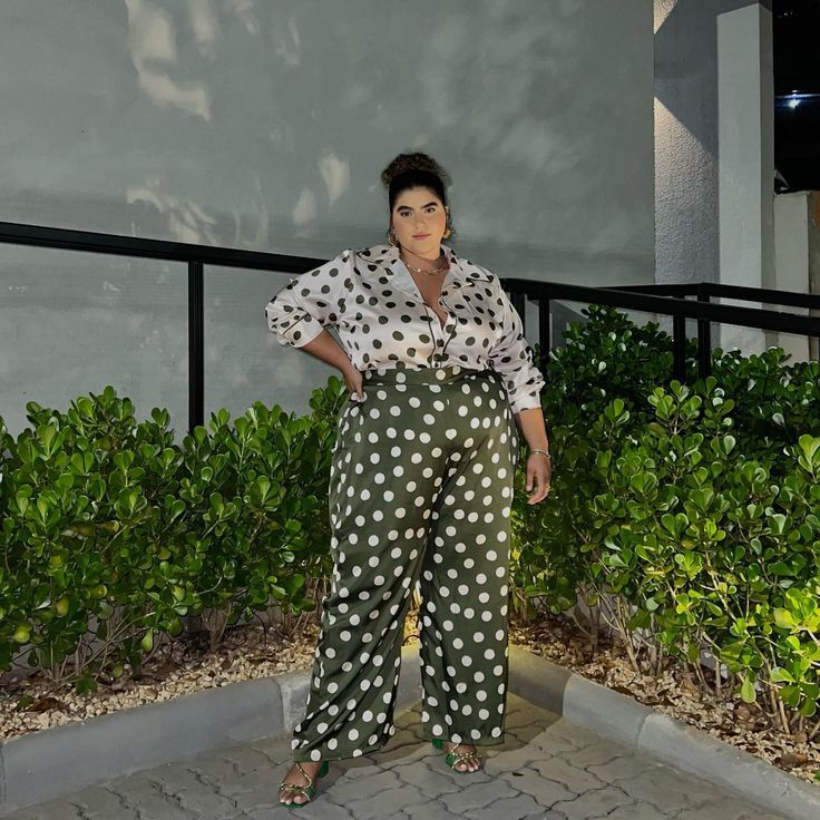 Mulher de pele morena e cabelo castanho preso em coque, sorri levemente, posando com a m&atilde;o na cintura. Ela veste camisa de bolinhas pretas em fundo branco e cal&ccedil;a pantalona verde-militar com bolinhas brancas, combinando com sand&aacute;lias verdes. Est&aacute; em um ambiente externo, com arbustos verdes e uma parede cinza ao fundo