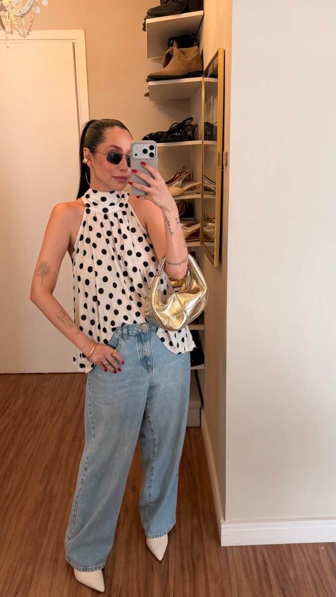 Mulher de cabelo escuro preso, &oacute;culos de sol e blusa branca de bolinhas pretas, jeans claro e sapatos brancos, posa para selfie no espelho. Ela segura uma bolsa dourada no bra&ccedil;o esquerdo. Ao fundo, prateleiras com sapatos e um lustre.