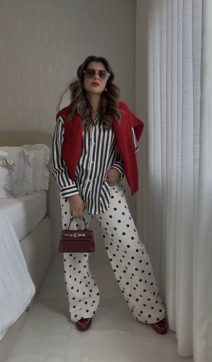 Mulher de &oacute;culos escuros, blusa listrada preta e branca, cal&ccedil;a pantalona branca de bolinhas pretas, su&eacute;ter vermelho nos ombros e bolsa vinho, posando em um quarto com cama e cortina branca