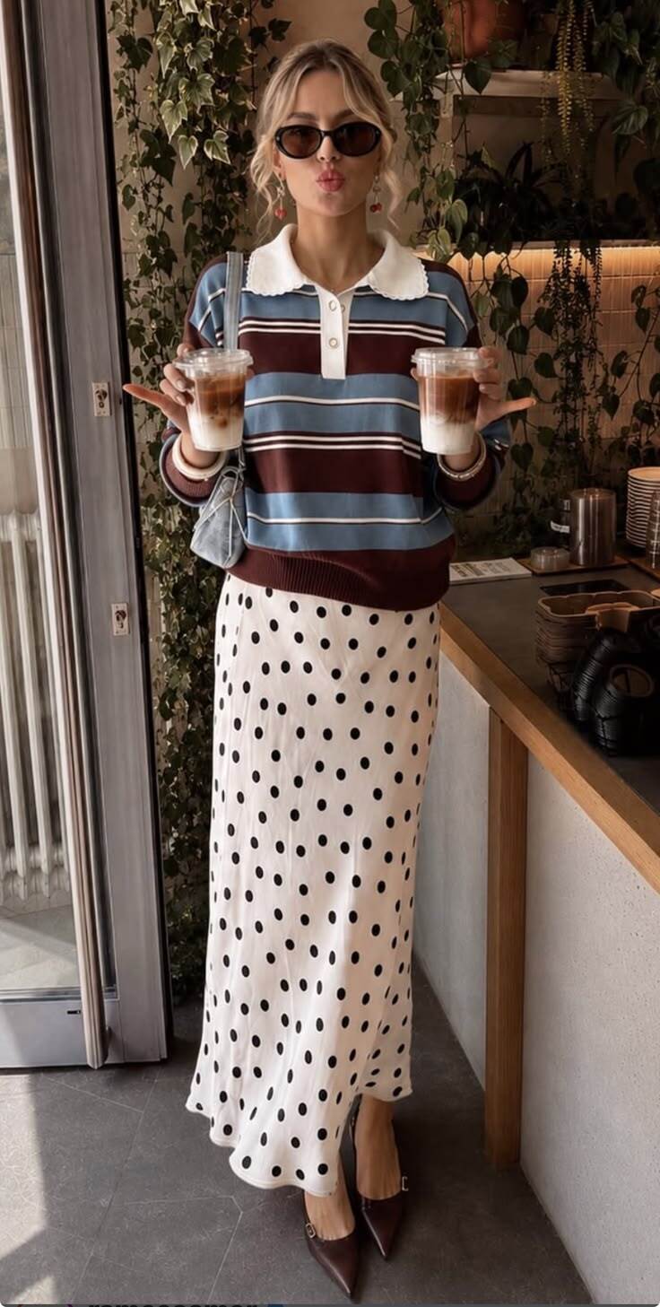Mulher loira de &oacute;culos escuros e batom vermelho, fazendo bico, segurando dois copos de caf&eacute; gelado. Ela veste um su&eacute;ter listrado azul, marrom e branco com gola polo, e uma saia longa branca de bolinhas pretas, em um ambiente interno com plantas penduradas