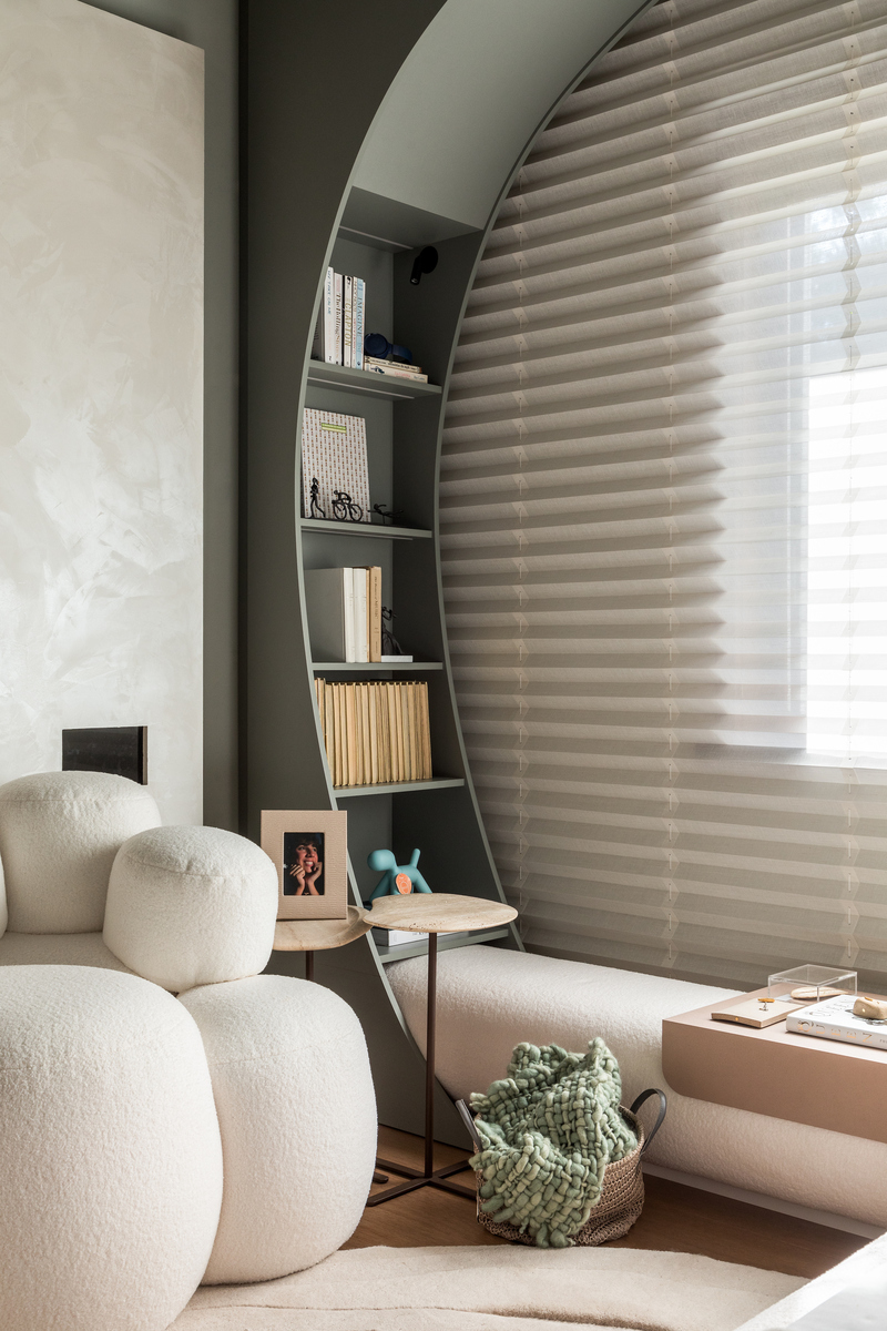 Ambiente interno com sof&aacute; modular branco, estante curva cinza-esverdeada com livros e objetos, e persiana horizontal bege cobrindo uma janela. H&aacute; duas mesas de cabeceira redondas, uma com porta-retrato e outra com um objeto azul, e uma cesta com manta verde no ch&atilde;o. Uma mesa de apoio rosa claro est&aacute; &agrave; direita