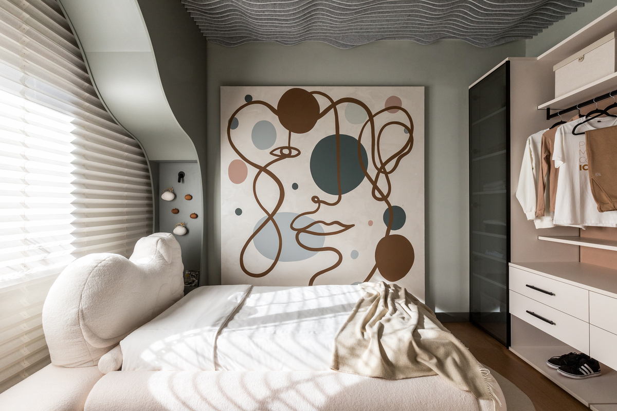 Quarto moderno com cama branca e cabeceira estofada, len&ccedil;&oacute;is brancos e manta bege. Na parede, um quadro abstrato com linhas marrons e c&iacute;rculos azuis e rosados. &Agrave; direita, um arm&aacute;rio aberto com prateleiras e roupas penduradas. &Agrave; esquerda, uma janela com persianas plissadas e uma parede curva com nichos decorativos. O teto tem um revestimento ondulado cinza.