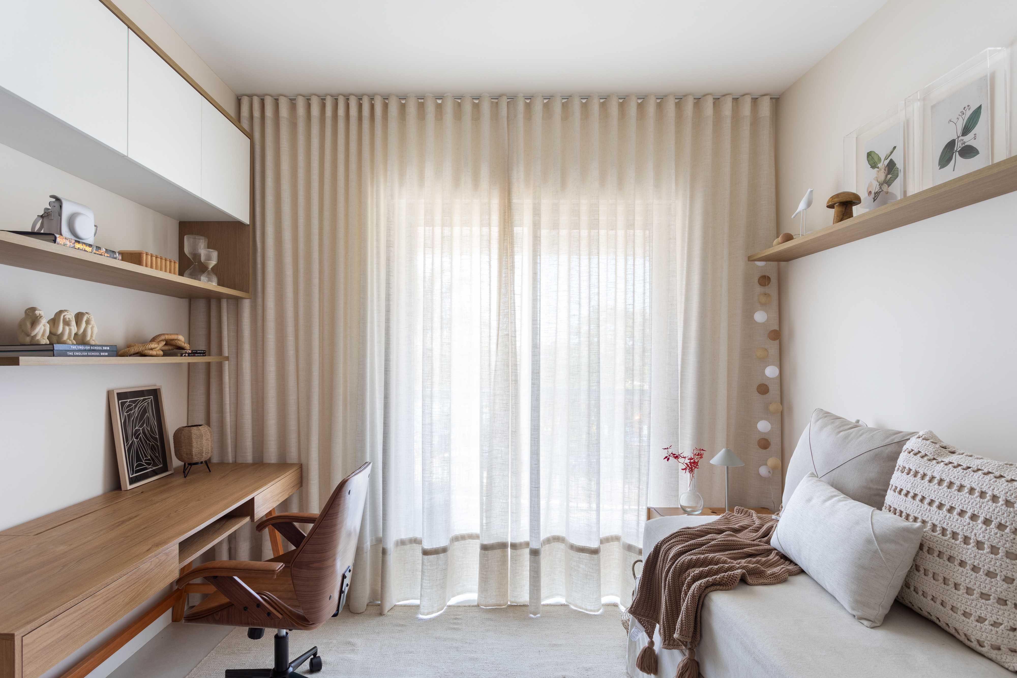 Quarto moderno com cama de solteiro &agrave; direita, coberta por manta marrom e almofadas claras. &Agrave; esquerda, escrivaninha de madeira clara com cadeira de escrit&oacute;rio marrom. Prateleiras e arm&aacute;rios brancos decoram a parede, e uma cortina bege transl&uacute;cida cobre a janela central, iluminando o ambiente