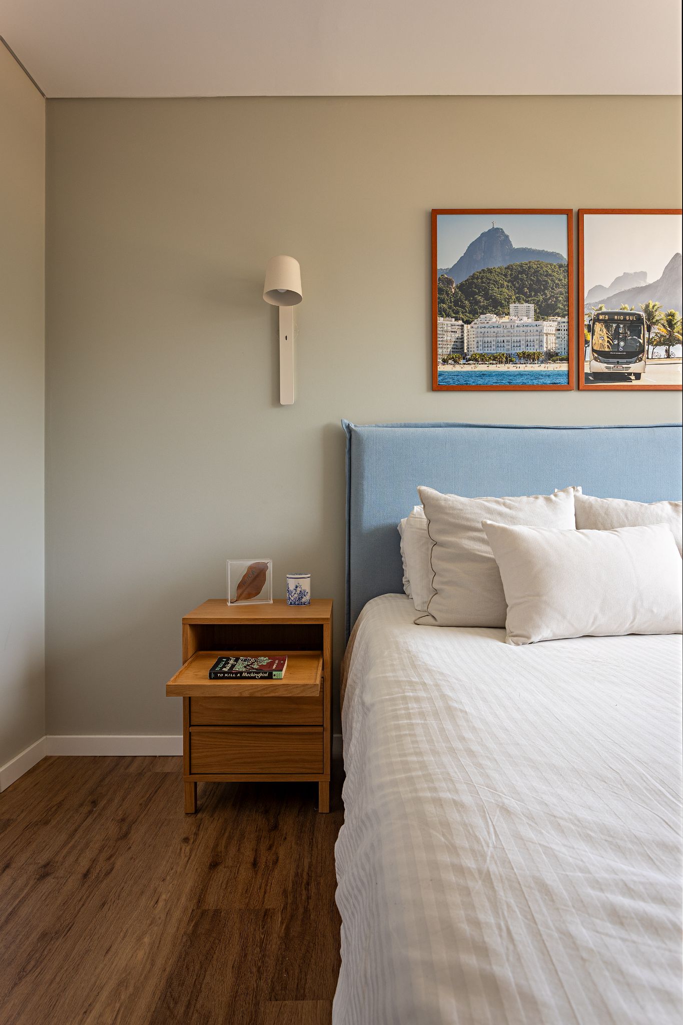 Quarto com parede verde-clara, cama com cabeceira azul e len&ccedil;&oacute;is brancos. &Agrave; esquerda, uma mesa de cabeceira de madeira com um livro, um copo decorado e uma folha seca em um quadro transparente. Acima da cama, dois quadros com paisagens do Rio de Janeiro, incluindo o Cristo Redentor e um &ocirc;nibus tur&iacute;stico. Uma lumin&aacute;ria de parede branca ilumina o ambiente. O ch&atilde;o &eacute; de madeira.