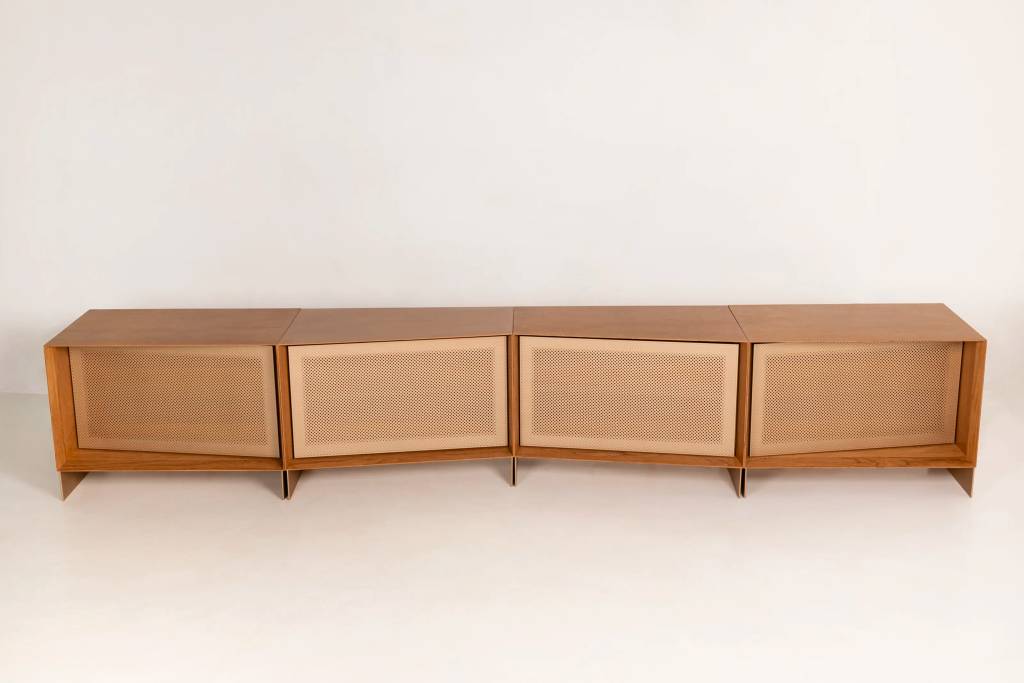 Um m&oacute;vel de madeira clara, estilo rack, composto por quatro m&oacute;dulos retangulares alinhados. Cada m&oacute;dulo possui uma porta frontal com tela perfurada bege, e os m&oacute;dulos centrais est&atilde;o ligeiramente angulados para dentro, criando um design curvo. O m&oacute;vel est&aacute; sobre um piso branco, com uma parede branca ao fundo