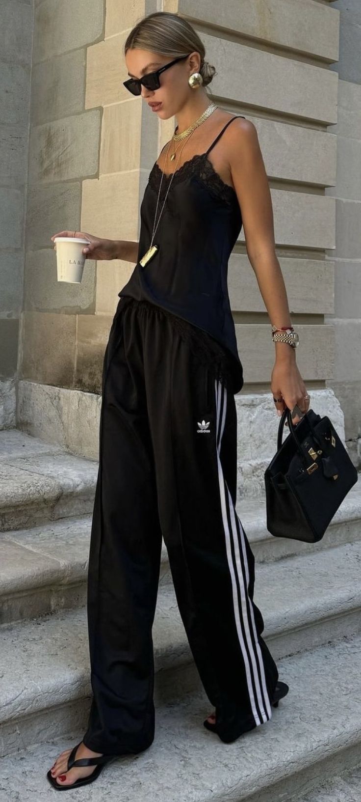 Mulher loira de &oacute;culos escuros e coque, vestindo top de al&ccedil;a preto com renda, cal&ccedil;a Adidas preta com listras brancas, chinelos pretos e unhas vermelhas. Ela segura um copo de caf&eacute; e uma bolsa preta, com joias douradas e rel&oacute;gio no pulso, em frente a uma parede de pedra e degraus