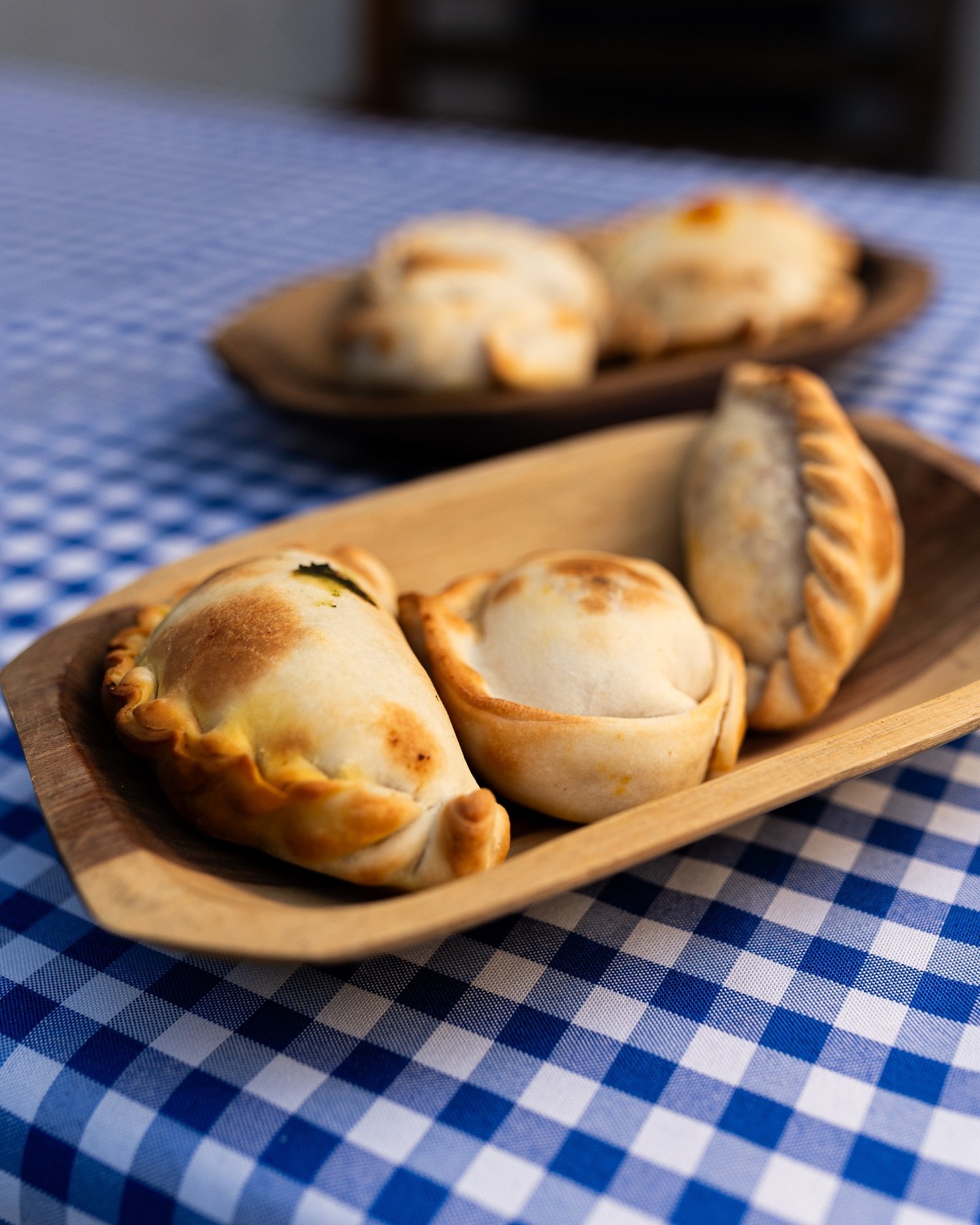 Tr&ecirc;s empanadas assadas em uma travessa de madeira clara sobre uma toalha de mesa xadrez azul e branca. Uma empanada tem recheio verde vis&iacute;vel, e outras duas travessas com empanadas desfocadas ao fundo