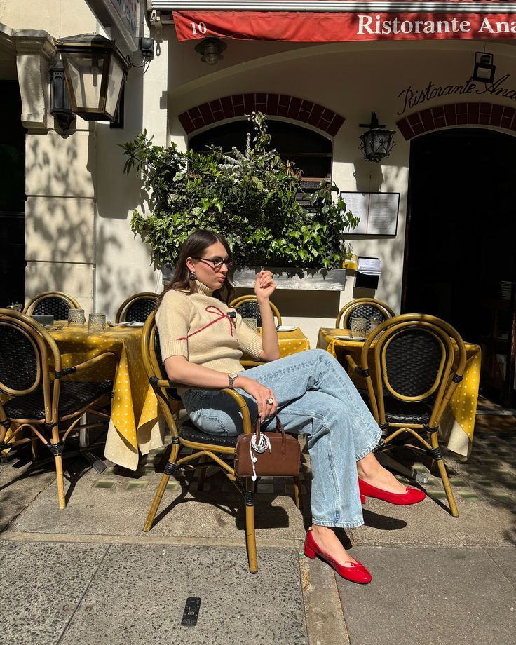 Mulher de &oacute;culos, su&eacute;ter bege e jeans claros sentada em cadeira de vime em cal&ccedil;ada, com sapatos vermelhos e bolsa marrom. Ao fundo, mesas de restaurante com toalhas amarelas e fachada com toldo vermelho e plantas.