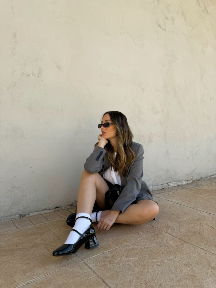 Mulher jovem de &oacute;culos escuros e blazer cinza sentada no ch&atilde;o de azulejos, com as pernas cruzadas e a m&atilde;o no queixo, olhando para a direita. Ela veste camiseta branca, shorts preto, meias brancas e sapatos Mary Jane pretos de salto bloco. O fundo &eacute; uma parede clara texturizada