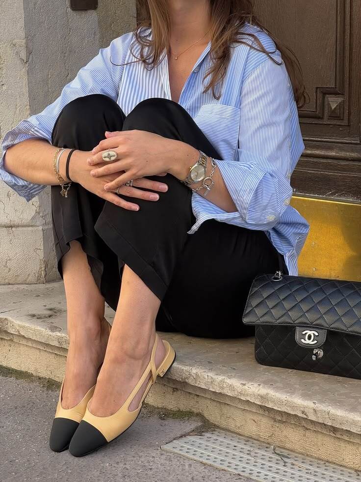 Mulher sentada em degraus de pedra, vestindo camisa listrada azul e branca, cal&ccedil;a preta, sapatilhas bege com biqueira preta e bolsa Chanel preta acolchoada ao lado. Ela usa rel&oacute;gio prateado, an&eacute;is e pulseiras nos bra&ccedil;os e m&atilde;os cruzadas sobre os joelhos