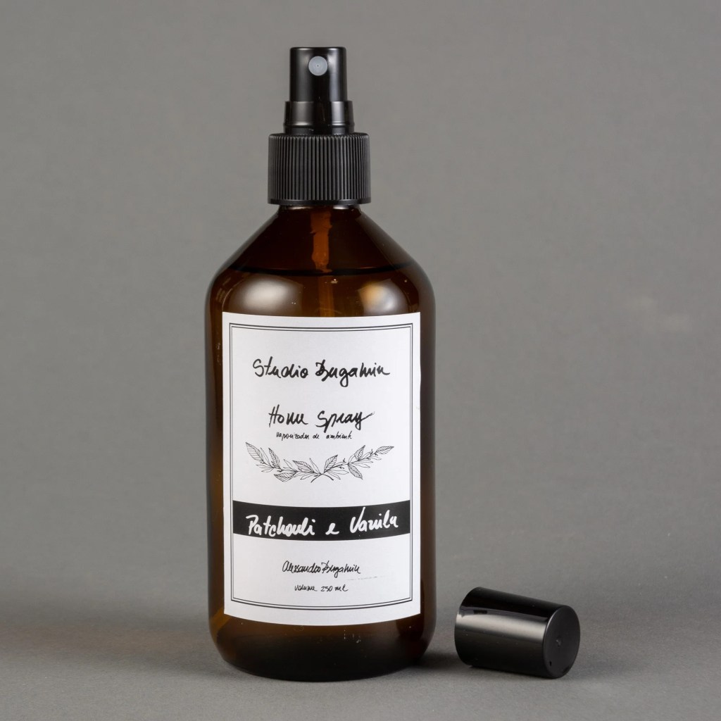 Frasco de vidro &acirc;mbar com borrifador preto, contendo Home Spray Patchouli e Vanila da Studio Bagamin, 250 ml, sobre fundo cinza. A tampa do borrifador est&aacute; ao lado