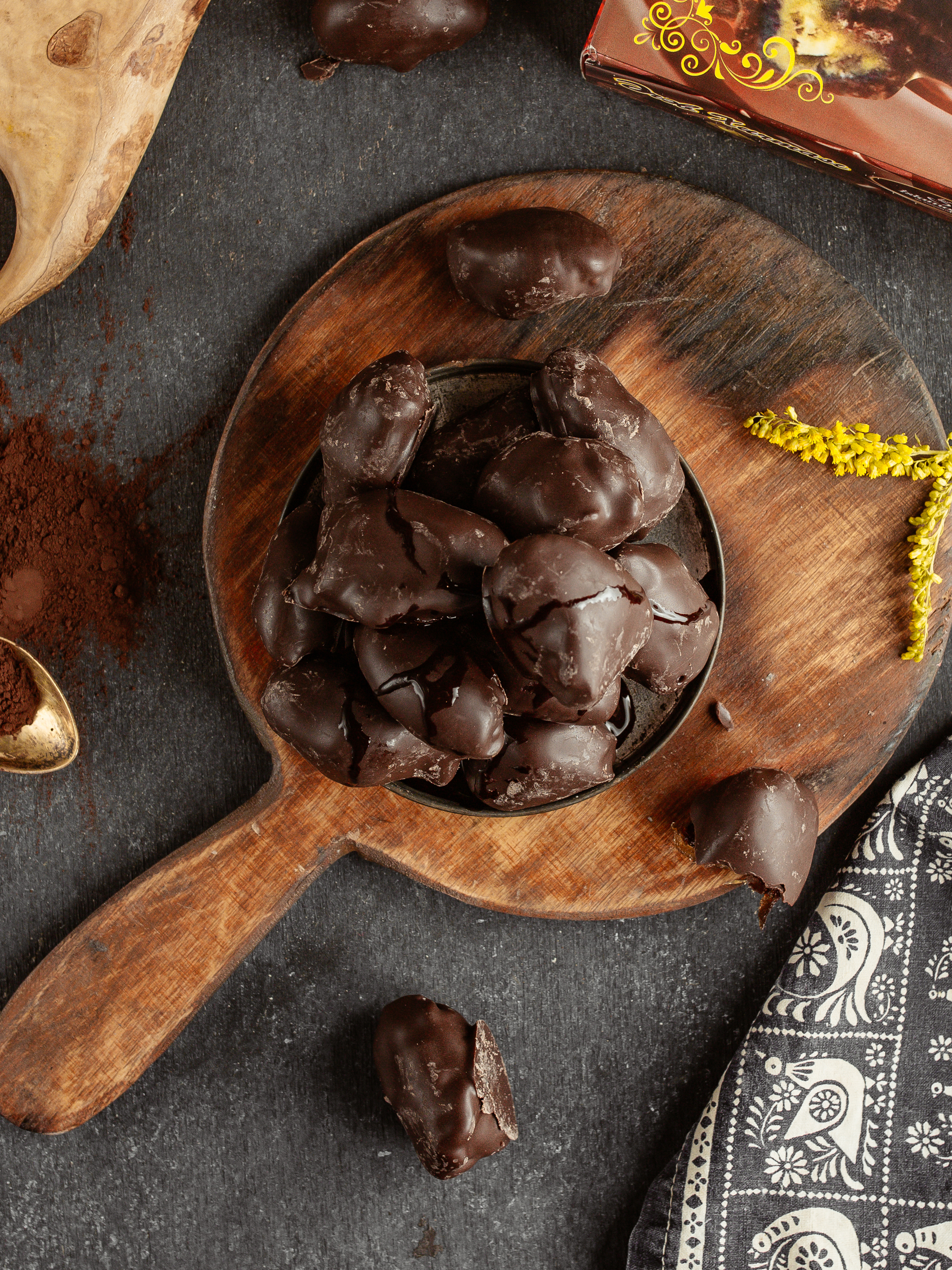 Doces de chocolate escuro em uma tigela preta sobre uma t&aacute;bua de madeira r&uacute;stica. H&aacute; p&oacute; de cacau e uma colher dourada &agrave; esquerda, um guardanapo estampado e um ramo de flores amarelas &agrave; direita, e uma caixa de doces ao fundo, tudo sobre uma superf&iacute;cie escura