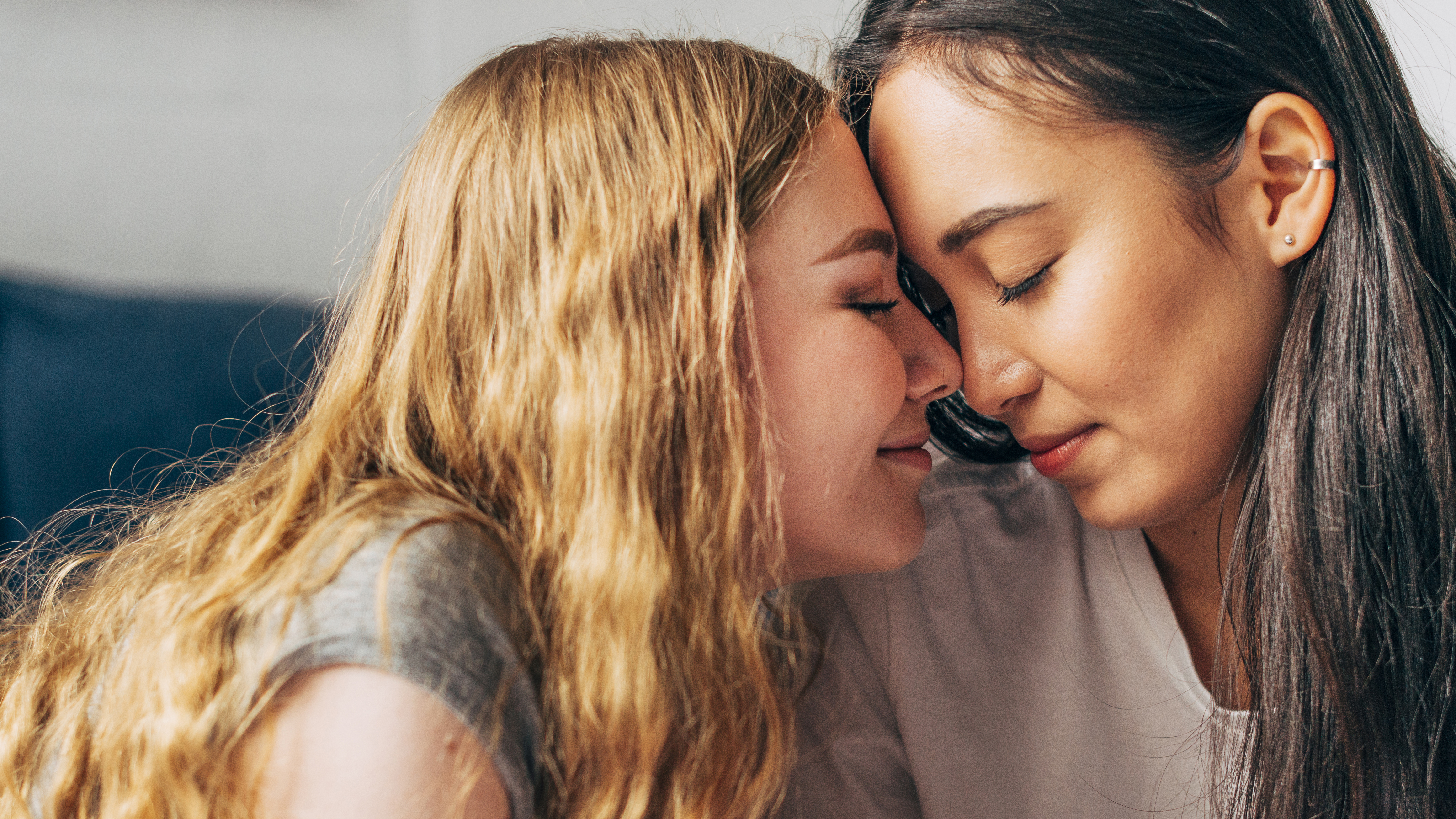 Duas mulheres, uma loira e uma morena, com os olhos fechados e rostos pr&oacute;ximos, testa com testa, em um momento de carinho e intimidade. A mulher loira veste uma camiseta cinza e a morena uma camiseta branca. O fundo &eacute; desfocado, com tons claros e um toque de azul.