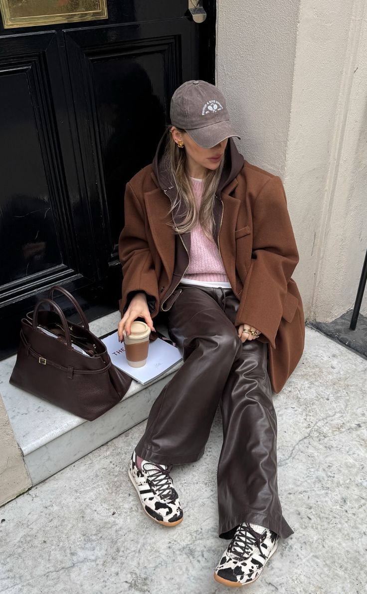Mulher sentada em degraus de pedra, vestindo bon&eacute; cinza, casaco marrom, moletom e su&eacute;ter rosa, cal&ccedil;a de couro marrom e t&ecirc;nis com estampa de vaca. Ela segura um copo de caf&eacute; reutiliz&aacute;vel, com uma bolsa marrom e uma revista ao lado