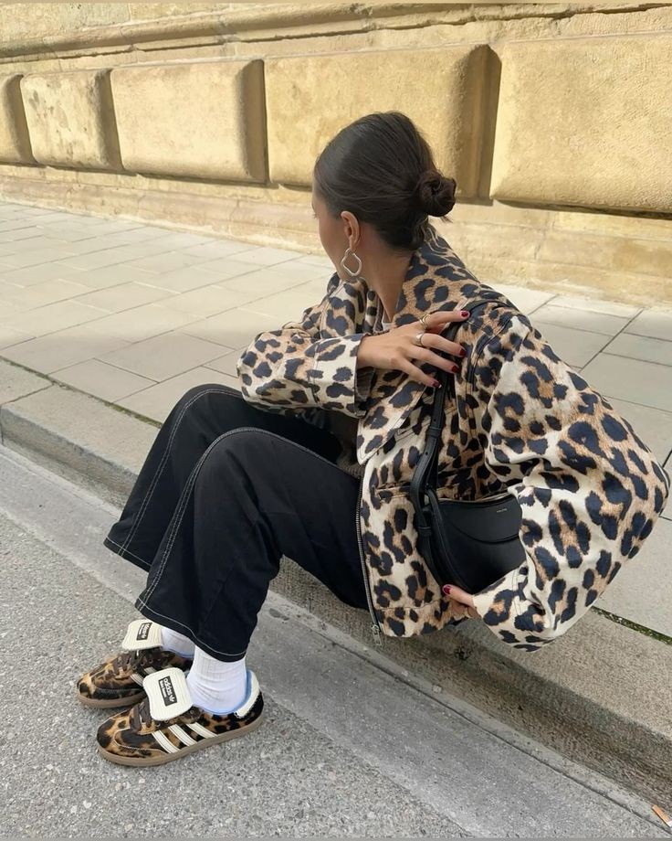 Pessoa sentada de costas, usando jaqueta e t&ecirc;nis com estampa de on&ccedil;a, cal&ccedil;a preta e bolsa preta, em uma cal&ccedil;ada com muro de pedra ao fundo
