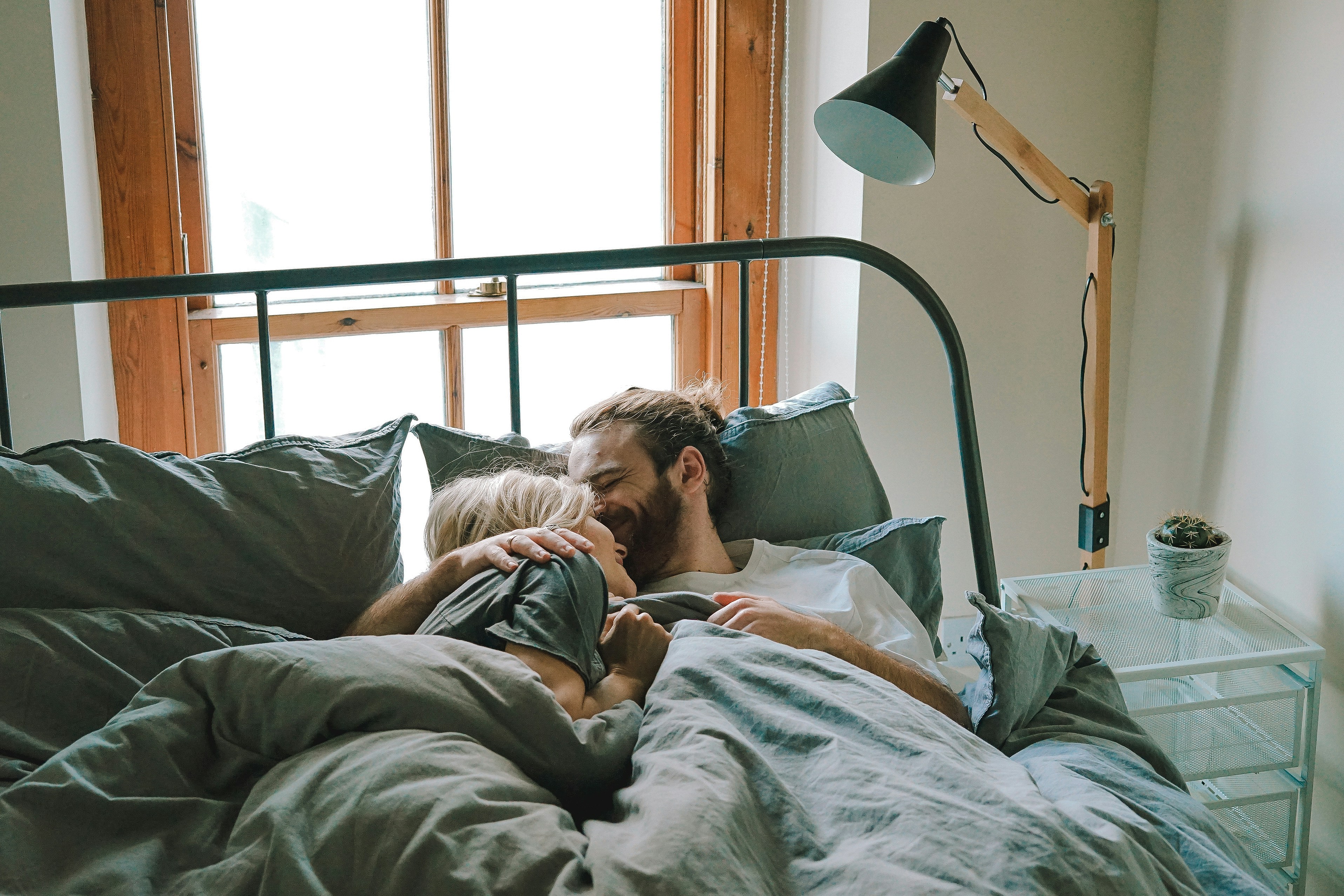 Casal jovem, um homem e uma mulher, deitados na cama sob um edredom cinza, sorrindo e se abra&ccedil;ando. A mulher tem cabelos loiros curtos e o homem barba e cabelo preso. Ao fundo, uma janela de madeira e uma lumin&aacute;ria de ch&atilde;o preta e de madeira. &Agrave; direita, uma mesa de cabeceira branca com um pequeno cacto.