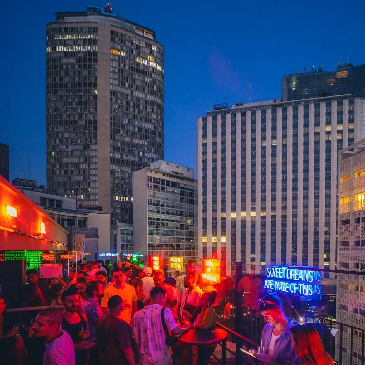 Pessoas em um bar movimentado no terra&ccedil;o de um pr&eacute;dio, com luzes neon vermelhas e verdes, e um letreiro azul Sweet Dreams Are Made Of This. Ao fundo, pr&eacute;dios altos iluminados contra o c&eacute;u azul escuro do anoitecer
