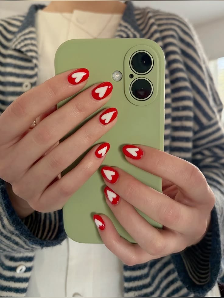M&atilde;os de uma pessoa com unhas ovais pintadas de vermelho com um cora&ccedil;&atilde;o branco no centro, segurando um celular verde-claro. A pessoa veste um casaco listrado azul e branco