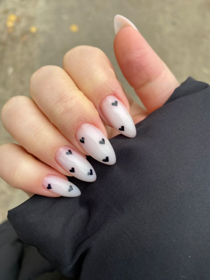 M&atilde;o feminina com unhas amendoadas longas, esmalte branco transl&uacute;cido e pequenos cora&ccedil;&otilde;es pretos. A m&atilde;o segura um tecido preto, com fundo desfocado