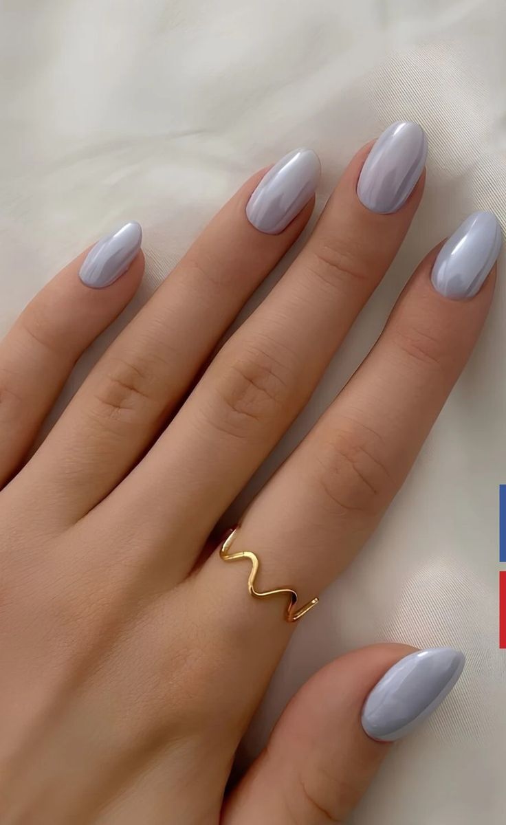 M&atilde;o feminina com unhas ovais pintadas de cinza-claro perolado, usando um anel dourado em formato de onda no dedo anelar, sobre um tecido branco amassado