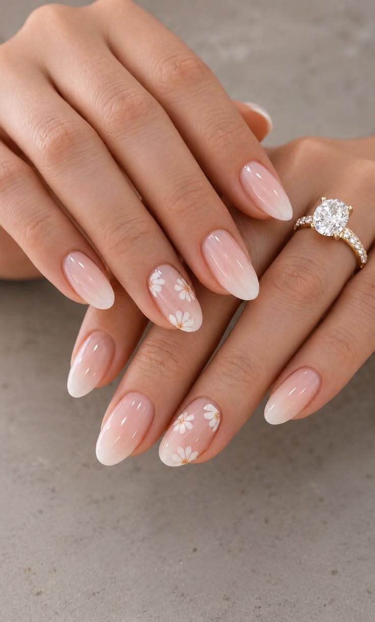 M&atilde;os femininas com unhas ovais em estilo francesinha degrad&ecirc; rosa claro para branco, com margaridas brancas e miolo dourado em algumas unhas. Um anel de noivado dourado com diamante grande e cravejado de brilhantes adorna o dedo anelar direito.