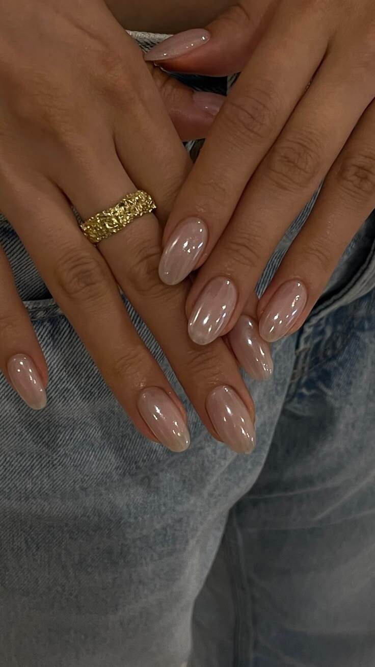M&atilde;os de uma pessoa com unhas ovais longas e esmalte perolado nude, usando um anel dourado texturizado no dedo anelar, sobre uma cal&ccedil;a jeans clara