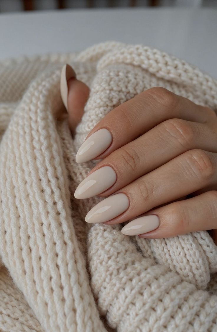 M&atilde;o feminina com unhas amendoadas em tom nude claro, segurando um tecido de tric&ocirc; creme com textura macia