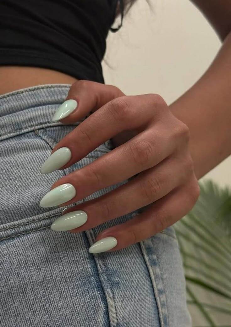 M&atilde;o feminina com unhas amendoadas pintadas de verde-&aacute;gua claro, tocando a cintura de uma cal&ccedil;a jeans azul-claro. Ao fundo, uma planta verde desfocada