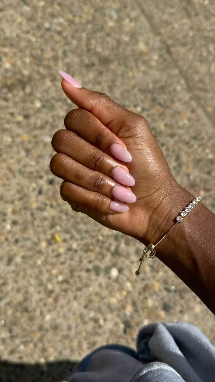M&atilde;o de pele escura com unhas amendoadas em tom rosa claro, usando uma pulseira dourada com pedras brilhantes no pulso, sobre um fundo de ch&atilde;o de pedras claras