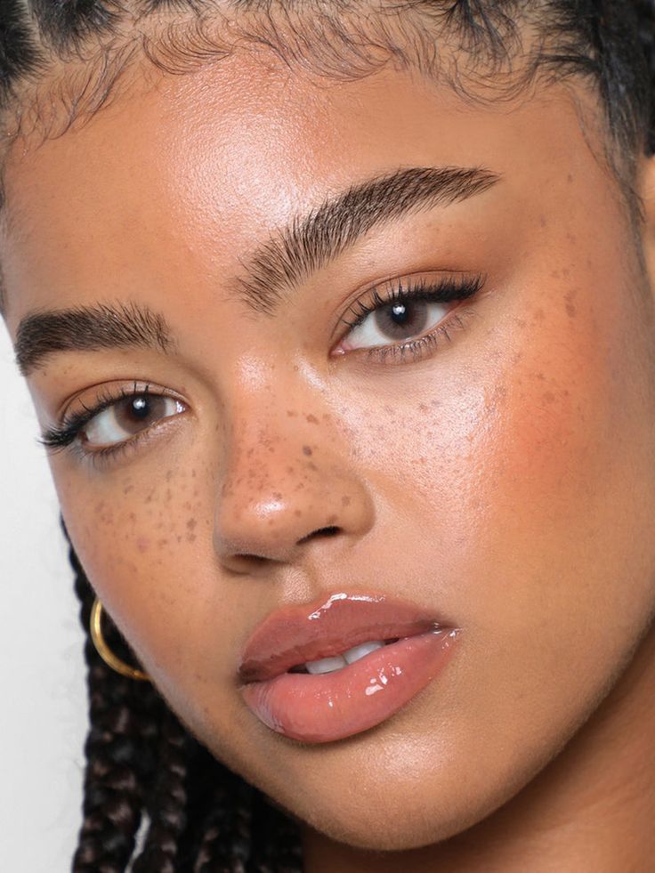 Rosto de uma mulher jovem com pele morena, sardas no nariz e bochechas, maquiagem natural com blush p&ecirc;ssego e l&aacute;bios com gloss nude. Ela tem olhos castanhos claros, sobrancelhas cheias e c&iacute;lios longos. Cabelo tran&ccedil;ado com baby hair na testa e argola dourada na orelha esquerda