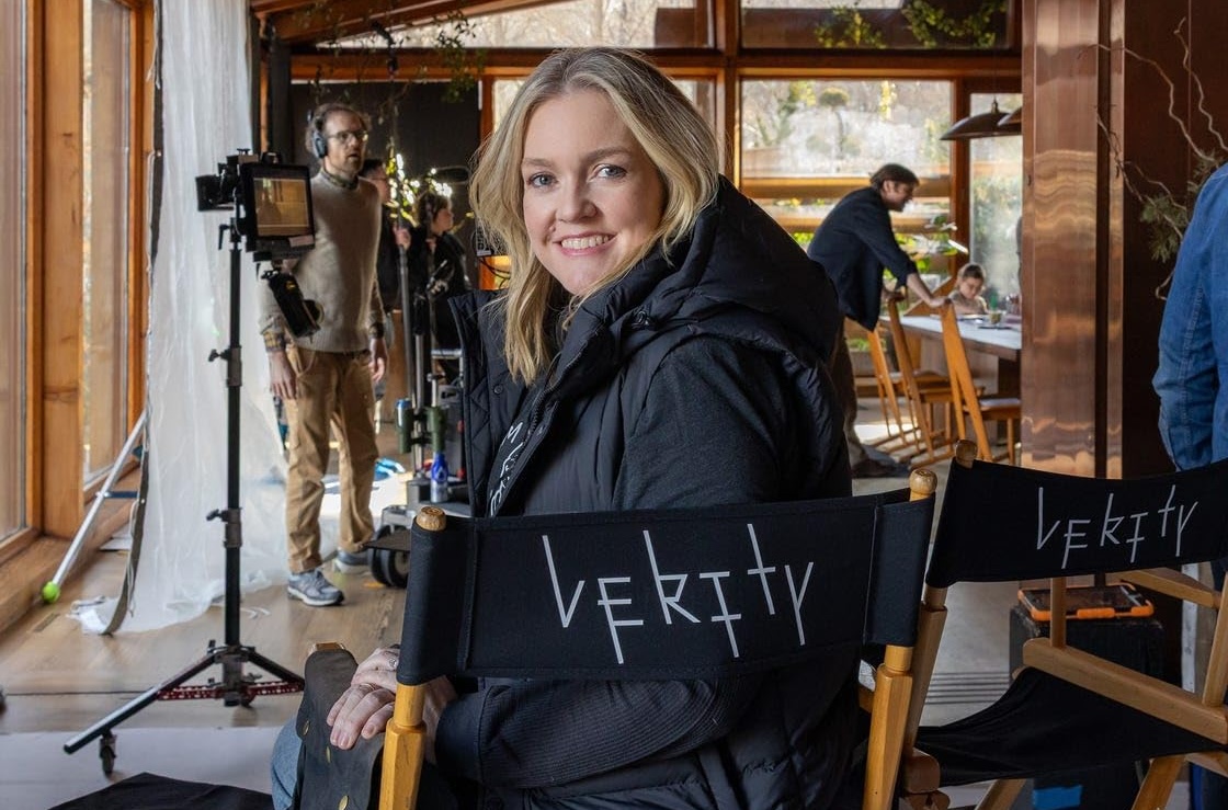Mulher loira de meia-idade, sorrindo, sentada em uma cadeira de diretor preta com Verity escrito, em um set de filmagem com equipamentos e equipe ao fundo