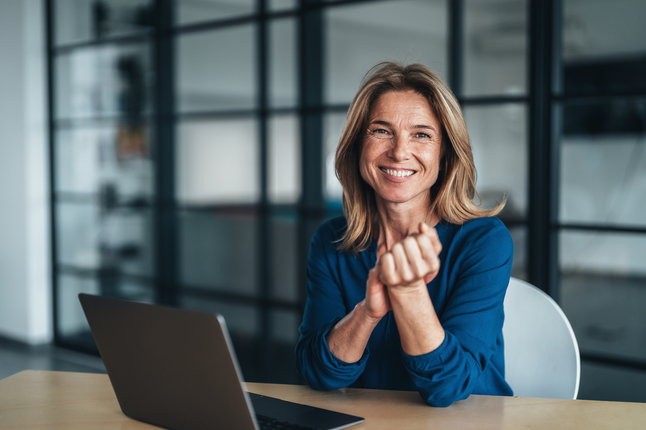 Mulher loira de meia-idade, sorrindo, com as mãos unidas sobre uma mesa de madeira clara, um notebook à esquerda e uma parede de vidro com divisórias escuras ao fundo