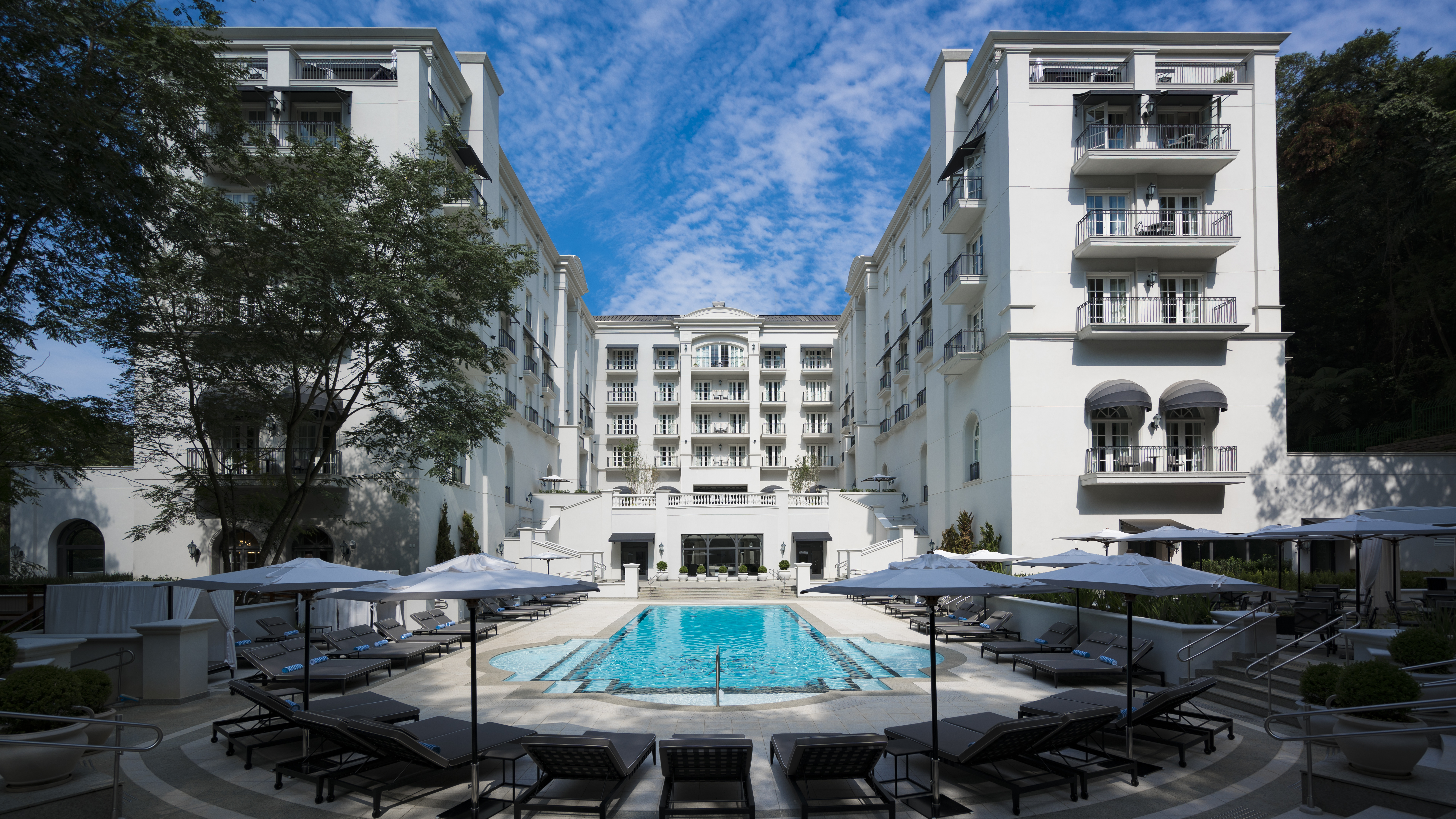 Piscina retangular de hotel luxuoso, cercada por espreguiçadeiras e guarda-sóis cinzas, com três edifícios brancos de arquitetura clássica ao fundo, sob céu azul com nuvens esparsas