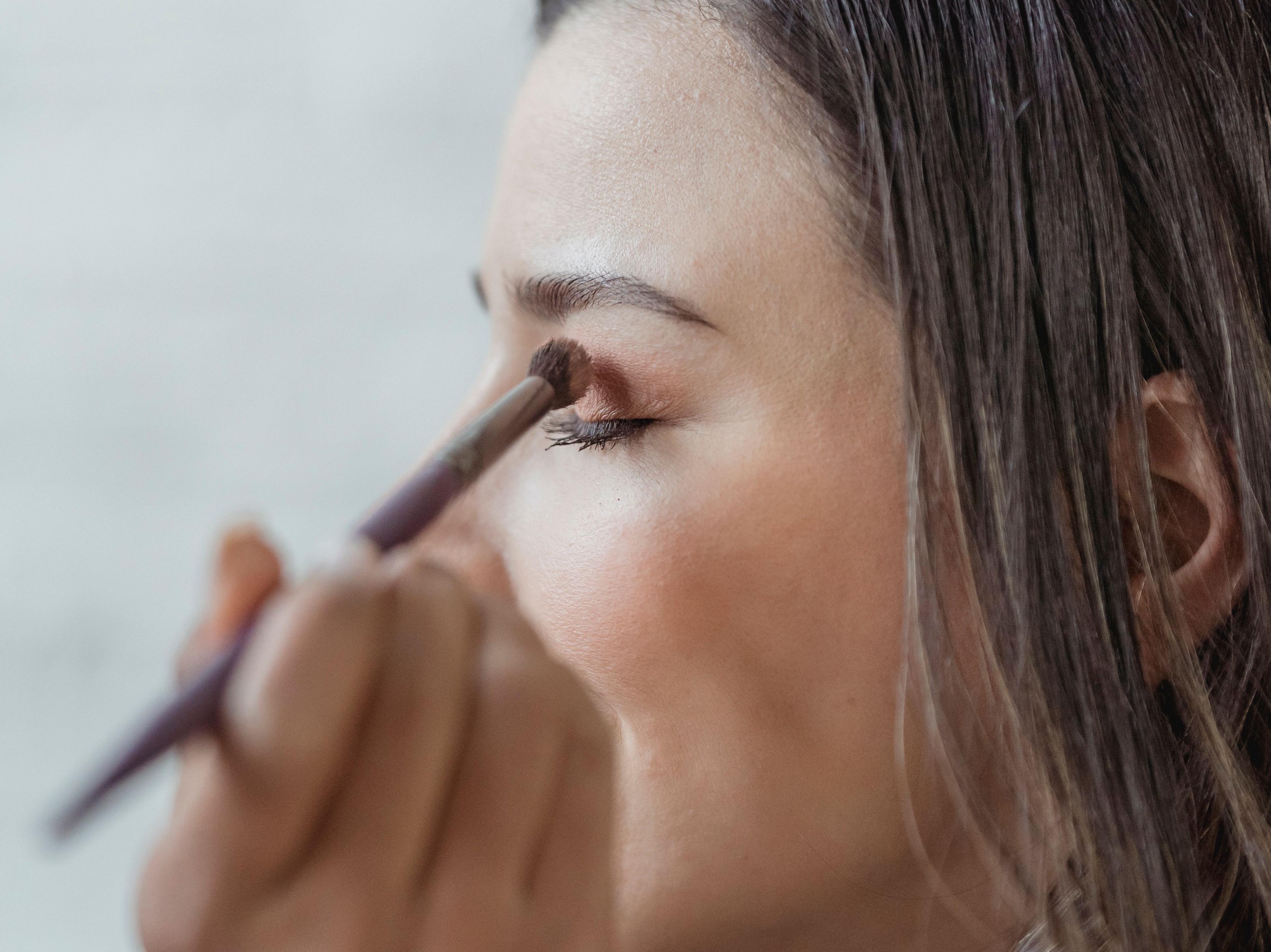 Mulher com cabelo castanho aplicando sombra marrom na pálpebra direita com um pincel de maquiagem, olhos fechados e expressão concentrada