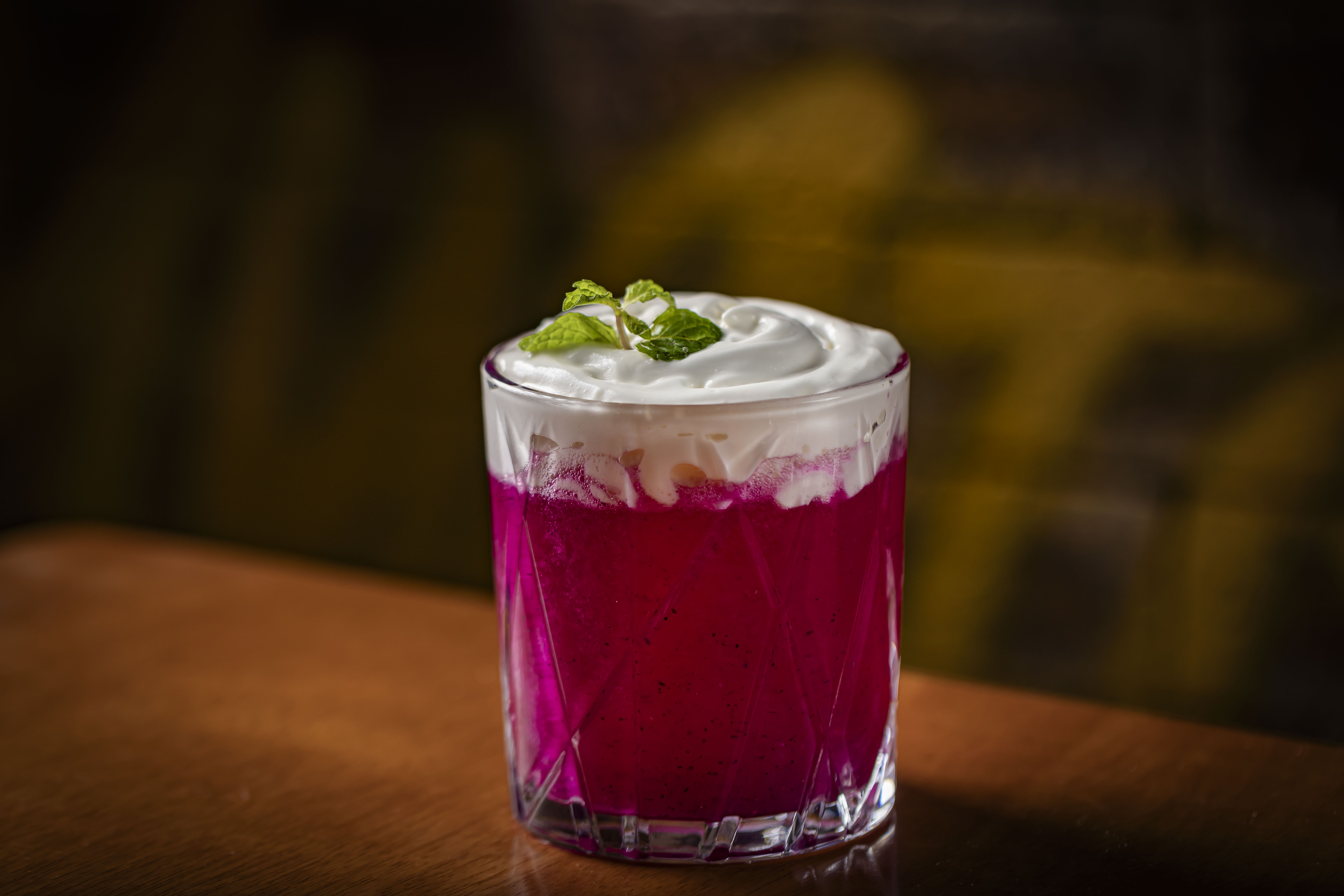 Copo de vidro com bebida rosa vibrante, coberta por espuma branca e folhas de hortelã, sobre uma mesa de madeira escura