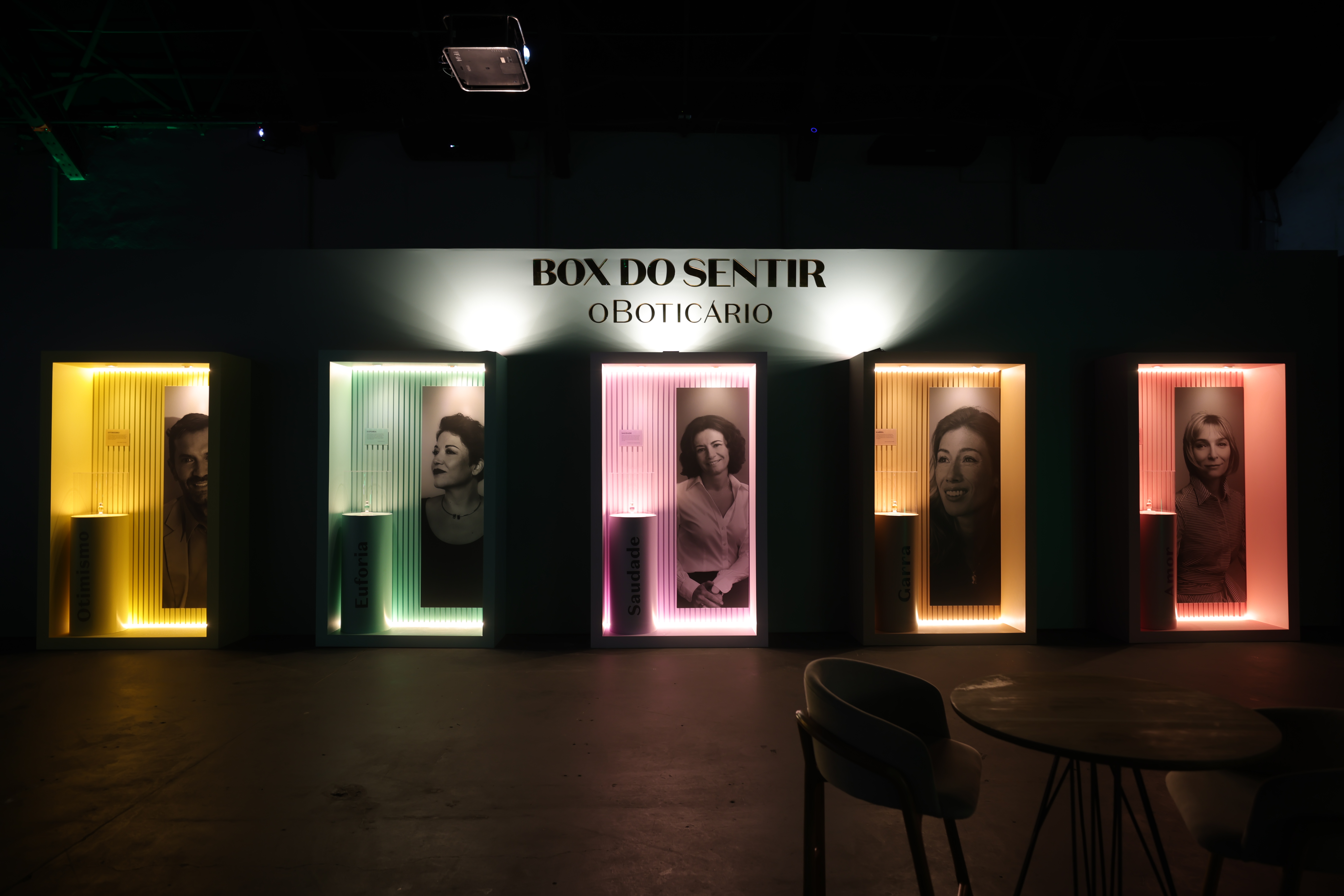 Exposição Box do Sentir O Boticário com cinco cabines iluminadas em tons vibrantes. Cada cabine exibe um retrato em preto e branco de uma pessoa e um cilindro com o nome de uma emoção, como Saudade e Euforia. O ambiente é escuro, com uma mesa e cadeiras à direita