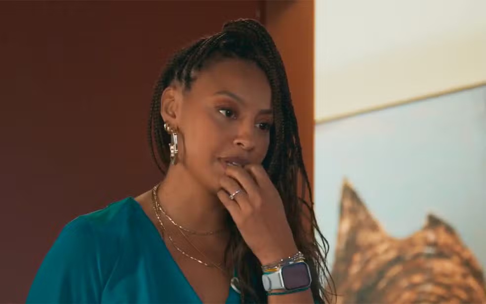 Mulher negra com tranças longas, vestindo blusa verde-água, colar dourado e brincos grandes, com expressão pensativa, mão direita no queixo, anel no dedo anelar e relógio inteligente no pulso esquerdo. Ao fundo, parede marrom e um quadro com imagem abstrata