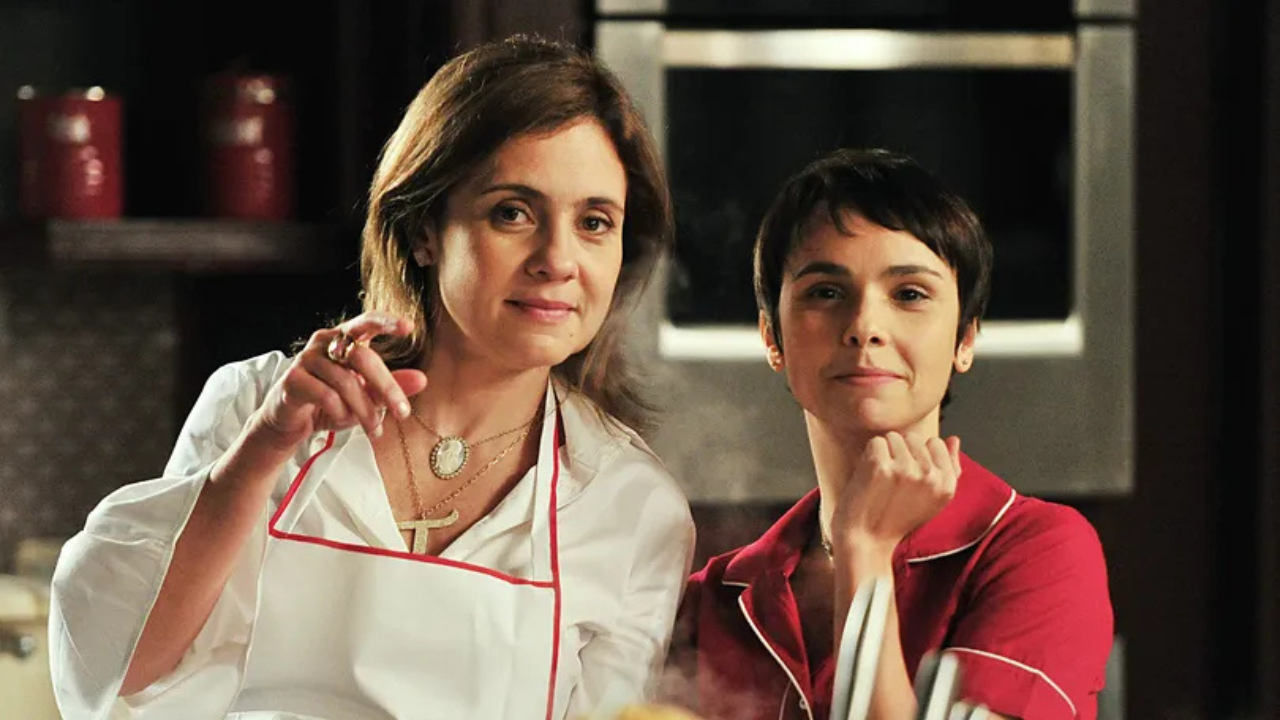Duas mulheres em uma cozinha. À esquerda, uma mulher de avental branco com borda vermelha e colar com pingente T aponta para baixo. À direita, outra mulher de cabelo curto e camisa vermelha com detalhes brancos apoia o queixo na mão, sorrindo levemente. Ambas olham para frente