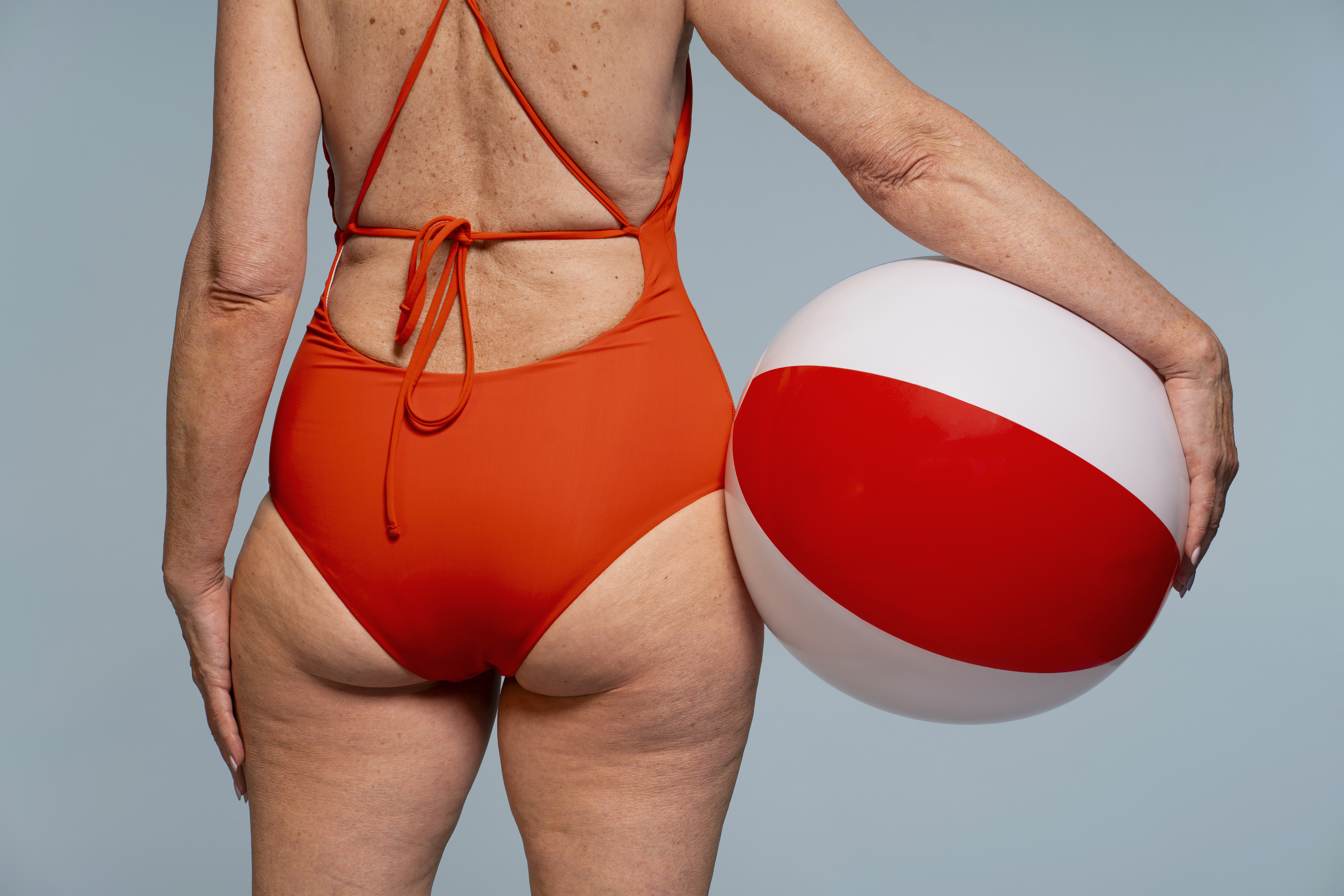 Mulher de costas, vestindo um maiô laranja com amarrações nas costas, segurando uma bola de praia branca e vermelha com a mão direita, em um fundo azul claro